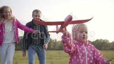 小可爱的金发女孩在秋天森林的背景下，带着粉红色的玩具飞机奔跑。 她的父亲和姐姐